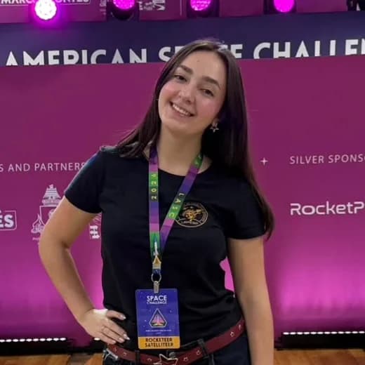 Maria Eduarda sorrindo, de camiseta preta e crachá, em frente ao painel do Latin American Space Challenge 2026